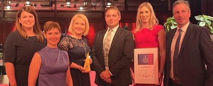 6 Personen stehen in Abendkleidung nebeneinander. Die zweite von rechts hält eine gerahmte Urkunde.